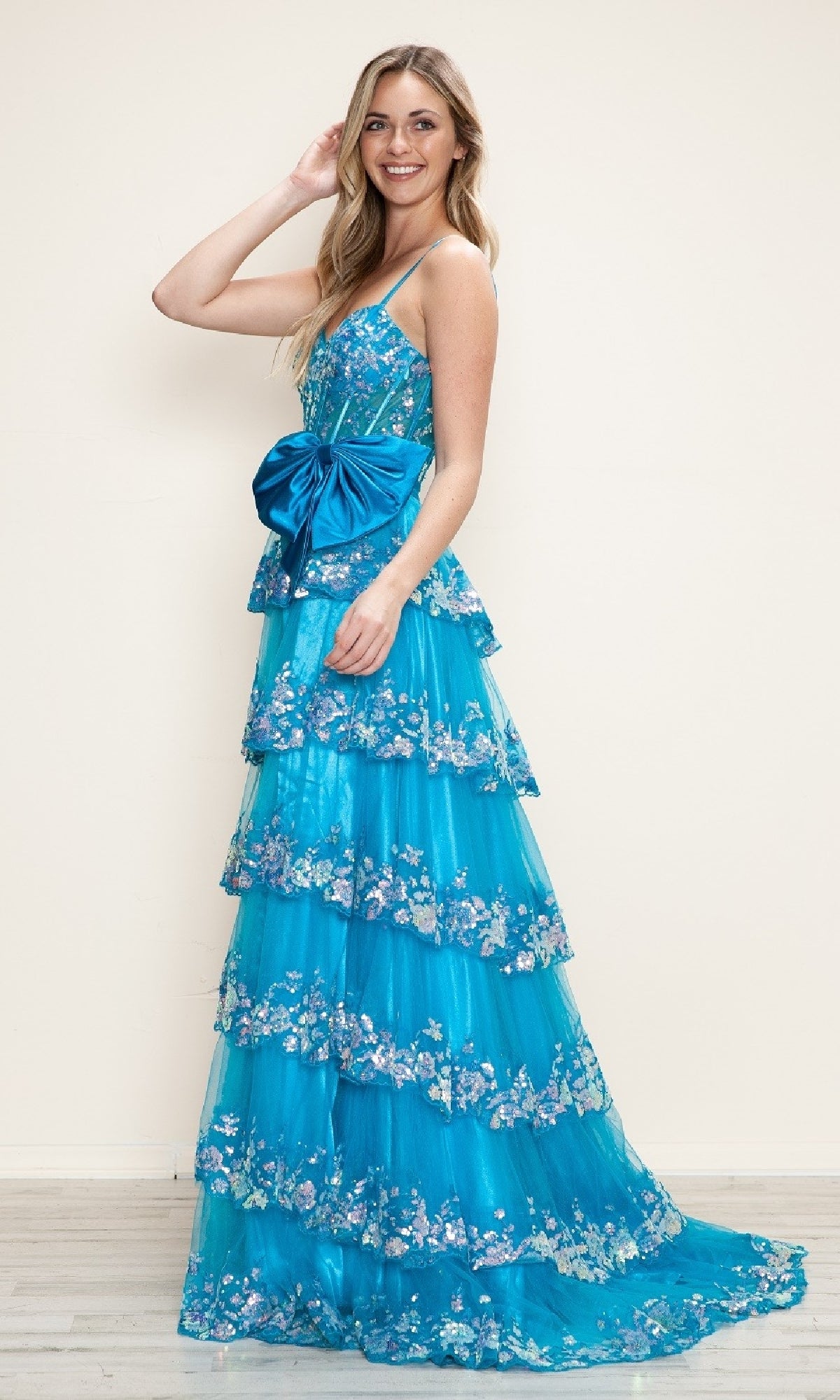 A woman smiles against a plain light background, wearing the Long Prom Dress: Poly USA 9626 in bright blue—an A-line gown with tiered skirt, spaghetti straps, and a large bow at the waist.